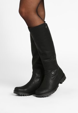 Bottes en cuir noires montantes jusqu'aux genoux portées avec des collants noirs transparents, présentant des semelles texturées et un petit logo sur le côté.