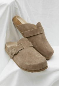 Suede brown clogs with a smooth texture, a rounded toe, and a decorative strap featuring a small gold-tone buckle. Cork footbed and rubber sole.