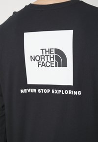 Schwarzes langärmliges T-Shirt mit einem weißen quadratischen Motiv, das das "THE NORTH FACE"-Logo und den Schriftzug "NEVER STOP EXPLORING" darunter zeigt.