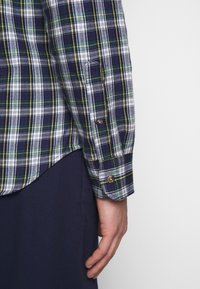 Main d’homme reposant à côté d’un pantalon bleu marine, portant une chemise à carreaux bleue, verte, blanche et jaune à manches longues avec poignets boutonnés.