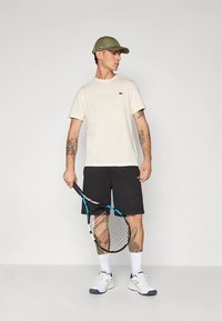 Herre tennisantrekk med en lys kremfarget t-skjorte, svarte shorts, hvite treningssko og en olivengrønn caps, holder en racket.