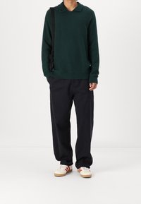 Dunkelgrüner, strukturierter Pullover mit Kragen, kombiniert mit lockeren, dunklen Hosen und weißen Sneakern mit roten Streifen. Schwarze Tasche über der Schulter.