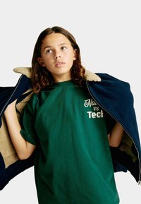 Ciemnoniebieska kurtka zapinana na zamek błyskawiczny z jasnobrązowym polarem w środku, noszona na zielonym podkoszulku z białym napisem: "Natura vs Technologia". Tkanina wydaje się być miękka.