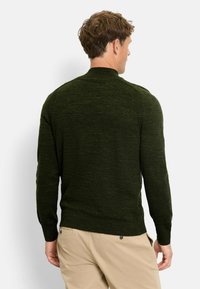 Dunkelgrüner Strickpullover mit hohem Kragen, langen Ärmeln und gerippten Bündchen sowie Saumdetails, kombiniert mit hellbeigen Hosen.