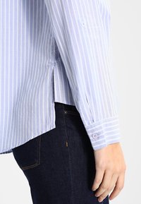 Hellblau-weiß gestreiftes Langarmhemd mit Knopfdetails an den Manschetten; der Stoff wirkt glatt und hat seitliche Schlitze für besseren Tragekomfort.