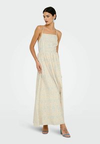 Robe maxi en tissu jaune clair avec imprimé floral bleu, dotée de fines bretelles, d'un corsage ajusté et d'une taille froncée. Associée à des talons bleus.
