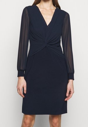 Navy blue knee-length dress with long sheer sleeves and a twisted knot detail at the front waist, worn by a standing person.