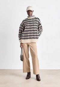 Gemusterter Pullover in Creme und Schwarz mit hohem Kragen, kombiniert mit beigen weiten Hosen und braunen Absatzstiefeln. Kleine Handtasche als Accessoire.