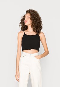 Top cropped preto com textura canelada e bainha em formato de ondas; combinado com calças brancas de cintura alta. A modelo está em pé com a mão no bolso.
