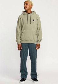 Olivengrøn hættetrøje med forlommer, snoretræk i hætten og lille logo. Kombineret med mørkeblå bukser og sorte slip-on sko.