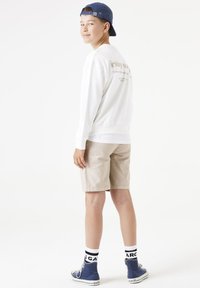 Sudadera blanca con texto en la parte posterior, pantalones cortos beiges, zapatillas deportivas azules y calcetas en blanco y negro. Cabello corto y gorra azul.