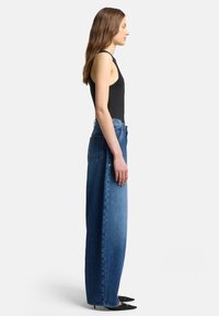High-waisted blue denim jeans with a wide-leg design, featuring back pockets and a raw hem. Worn with a fitted black tank top and pointed shoes.
