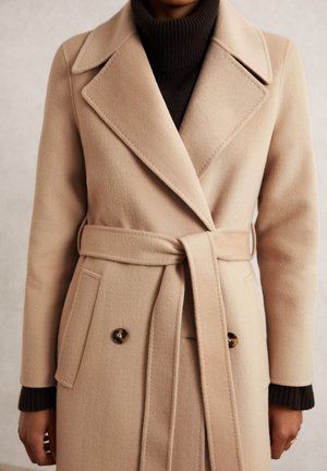 Beige uldfrakke med bælte og brede slag, båret over en mørkebrun ribbet rullekravesweater.