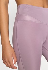 Vysoké legíny vo svetlofialovej farbe, vyrobené z hladkej, elastickej tkaniny s plochými švami a tónovaným farebným blokovaním na páse.