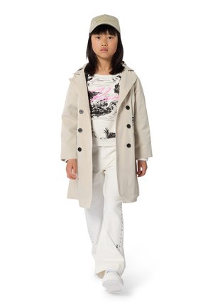 Beige trenchcoat met zwarte knopen, een grafisch zwart-wit shirt met roze accenten, witte broek met studs en een lichtkleurige pet.