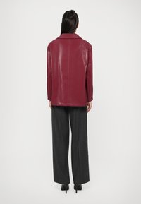 Giacca in pelle bordeaux con colletto, maniche lunghe e vestibilità dritta. Indossata sopra pantaloni grigi a gamba larga, mostrando una texture lucida.