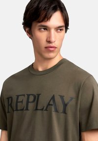 Oliven grøn bomulds T-shirt med et stort sort "REPLAY" grafik på brystet. Kortærmede og med rund hals.
