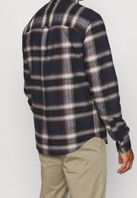 Koszula flanelowa w kratę w kolorze granatowym, szarym i burgundowym; długie rękawy z guzikami oraz zaokrąglony dół; miękka tekstura materiału.