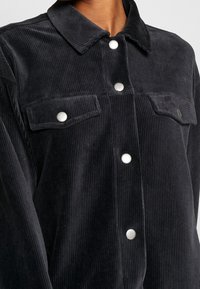 Camisa de pana negra con cuello clásico, botones a presión y dos bolsillos en el pecho. Presenta canalones verticales para aportar textura y profundidad.