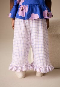 Svetloružové kárované nohavice s volánikovým lemom, skombinované s bielymi teniskami. Tkanina pôsobí mäkko a má textúru, vhodná na bežné nosenie.