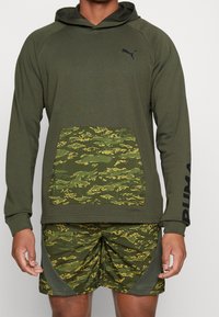 Olivgrüner Kapuzenpullover mit einer camouflagemusternden Tasche. Kombiniert mit camo Shorts, die gelbe Akzente und einen strukturierten Stoff aufweisen.