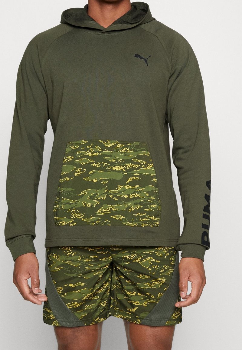 Olivgrüner Kapuzenpullover mit einer camouflagemusternden Tasche. Kombiniert mit camo Shorts, die gelbe Akzente und einen strukturierten Stoff aufweisen.