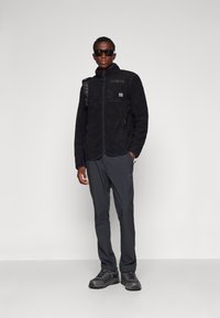 Svart fleecejacka med full zip, texturerad yta, sidofickor och en logotypapplikation; kombinerad med mörka byxor och robusta skor.