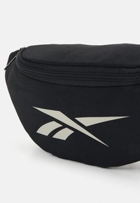 Sac banane noir en nylon, de forme arrondie, avec une fermeture à glissière sur le dessus et un logo gris clair bien visible.