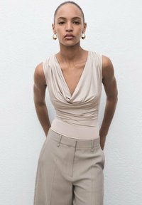 Top sin mangas beige con escote drapeado, combinado con pantalones beige de cintura alta. Textura de tela suave, diseño simple con acentos mínimos.