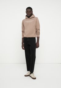 Felpa in pile marrone con logo frontale, abbinata a jeans neri e sneakers crema. L'outfit presenta una vestibilità rilassata e maniche lunghe.