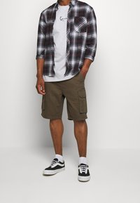 Camisa a cuadros en negro, gris y rojo, sobre una camiseta gris clara, combinada con pantalones cortos de cargo verde oliva y zapatillas negras.