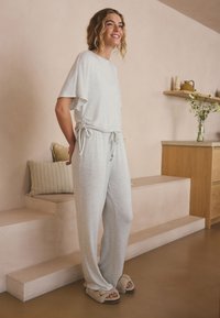 Jeune femme en tenue d’intérieur gris clair et chaussons duveteux, debout et souriante dans un espace de vie minimaliste aux tons neutres.