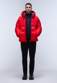 Rote Pufferjacke mit Kapuze, innen navy und mit Reißverschlusstaschen. Schwarze Hose und Stiefel runden den Look ab.