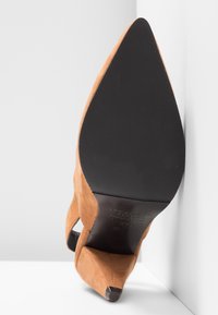 Chaussure à talon haut en daim marron avec un bout pointu, une semelle noire et des découpes sur les côtés, présentant une finition texturée.