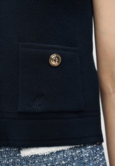 Μπλε υφασμάτινο γιλέκο με υφή, με τετράγωνη τσέπη που έχει ένα χρυσό κουμπί και κεντημένο γράμμα "Μ" πάνω από μπλε και λευκό παντελόνι με υφή.