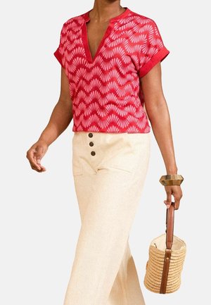 Vrouw die een rood topje met patroon en beige, hooggetailleerde broek draagt, en een gevlochten handtas met een bruine leren hengsel vasthoudt.