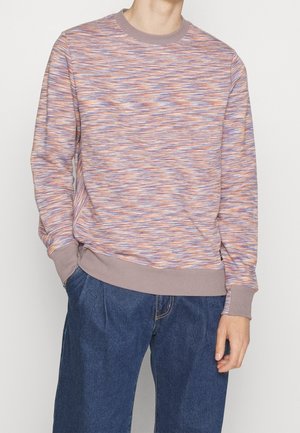 Man draagt een pastelgekleurde sweater met meerdere kleuren en een patroon, met licht mauve mouwen en halslijn, gecombineerd met losse donkerblauwe spijkerbroek, handen ontspannen.