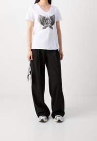 Vit kortärmad T-shirt med en svart och silver grafisk design av vingar och ett öga. Matcher med lösa svarta byxor och reflekterande sneakers.