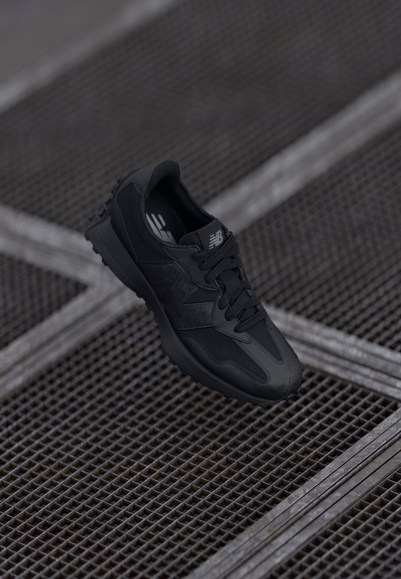 Baskets noires New Balance avec lacets reposant sur une surface en grille métallique avec des barres métalliques diagonales en dessous.