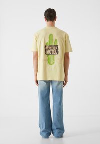 Geltona medvilninė marškinė su kaktuso grafika ir užrašu "Western Motel" nugarinėje pusėje, derinama su šviesiai mėlynomis plačiomis džinsinėmis kelnėmis.