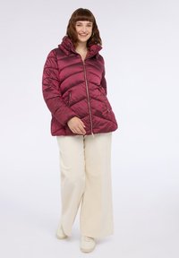 Burgunderfarbene Steppjacke mit hohem Kragen, diagonal gestepptem Muster, zwei seitlichen Taschen und einem Reißverschluss, getragen über cremefarbenen weiten Hosen.