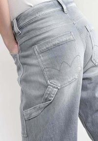 Los jeans de denim gris claro presentan una cintura alta, múltiples bolsillos y un distintivo bordado en los bolsillos traseros. El material está ligeramente desgastado.
