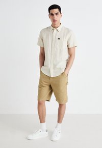 Kortermet, lys beige stripete skjorte med brystlomme, kombinert med brune shorts og hvite sneakers. Rent design, uformell stil.