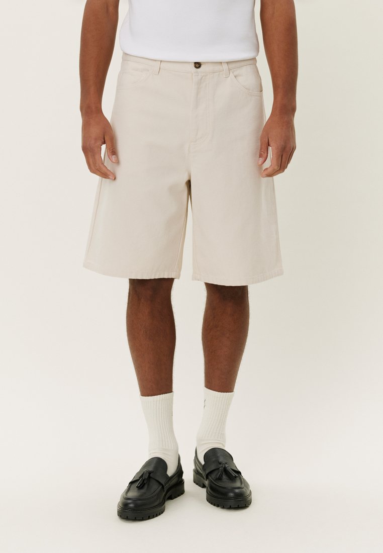 Shorts en coton beige à coupe droite, dotés de poches latérales, d'une fermeture à bouton et d'une texture lisse. Portés avec des chaussettes blanches et des chaussures noires.