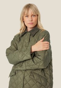 Chaqueta verde acolchada con cuello de pana, que cuenta con botones a presión y un bolsillo con parche, mostrando un patrón texturizado en forma de diamante.