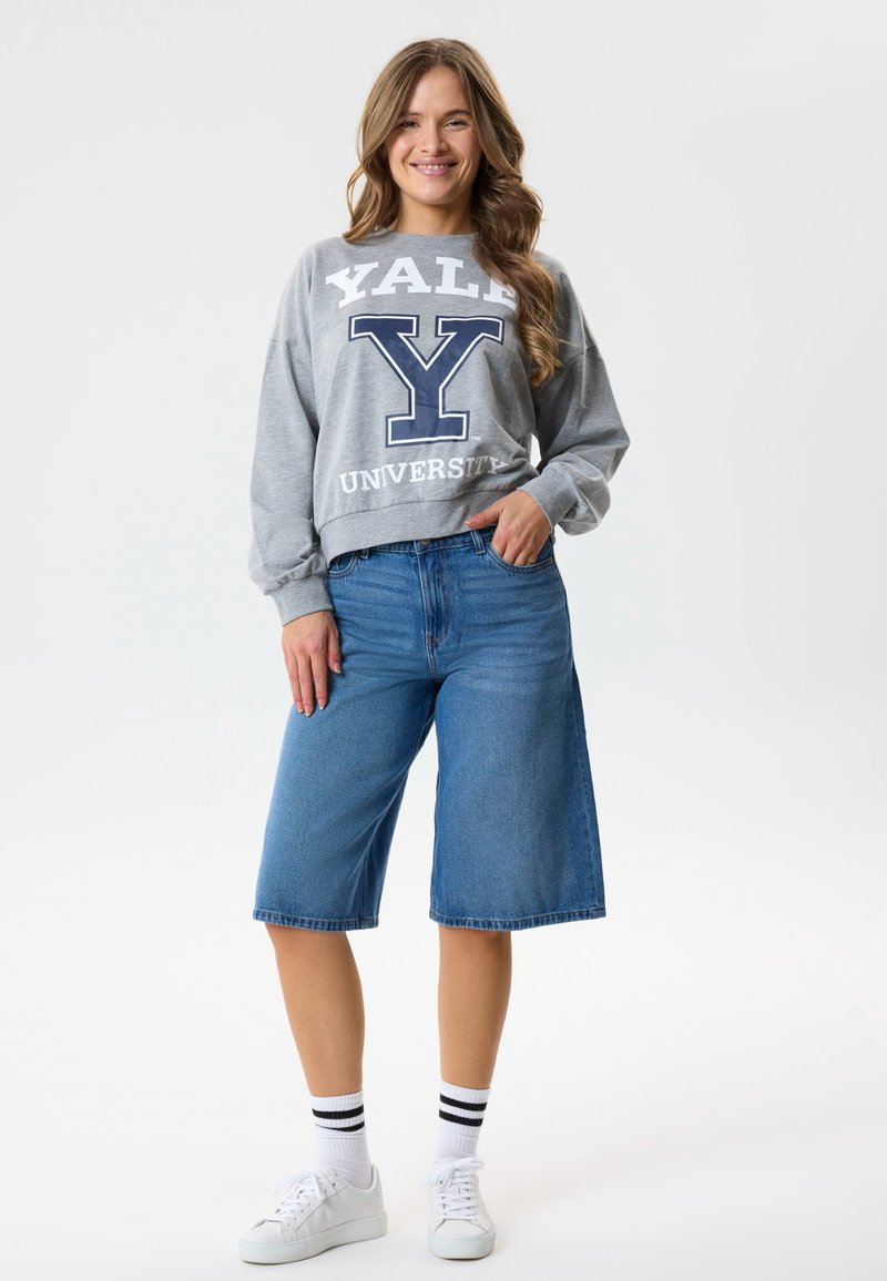 Femme souriante posant avec un sweat-shirt gris de l'université de Yale, un short en jean bleu jusqu'aux genoux, des baskets blanches et des chaussettes blanches à rayures noires.