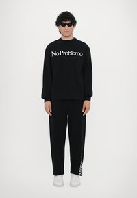 Svart hettegenser med teksten "No Problemo" i hvitt. Svart joggebukse med sideprint. Modellen bruker mørke solbriller og hvite joggesko.
