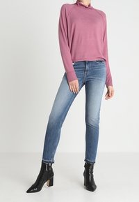 Rosa langärmliger Pullover mit Stehkragen, kombiniert mit blauen Slim-Fit-Jeans und schwarzen Stiefeletten mit quadratischem Absatz und Fransendetails.
