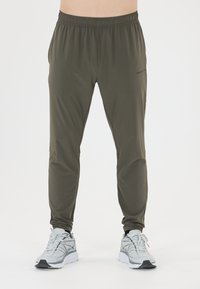 Man in olijfgroene sportbroek en witte hardloopschoenen, staande tegen een effen witte achtergrond.