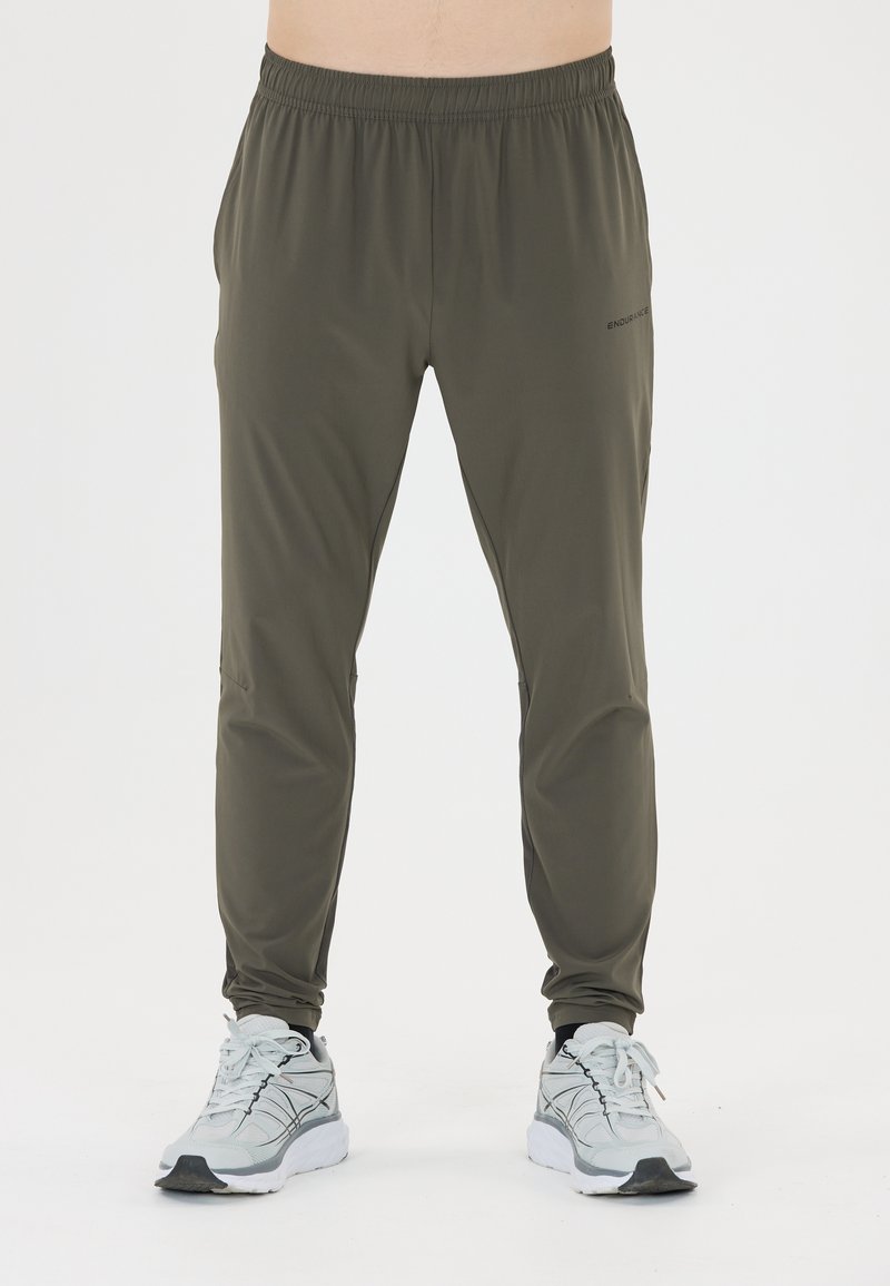 Man in olijfgroene sportbroek en witte hardloopschoenen, staande tegen een effen witte achtergrond.